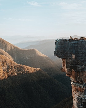 Scenic mountain landscape with a rocky cliff edge overlooking rolling hills at sunrise.