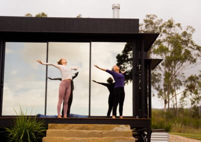 Two women practicing yoga with arms raised on a cabin deck surrounded by bushland at The Overflow 1895, Scenic Rim